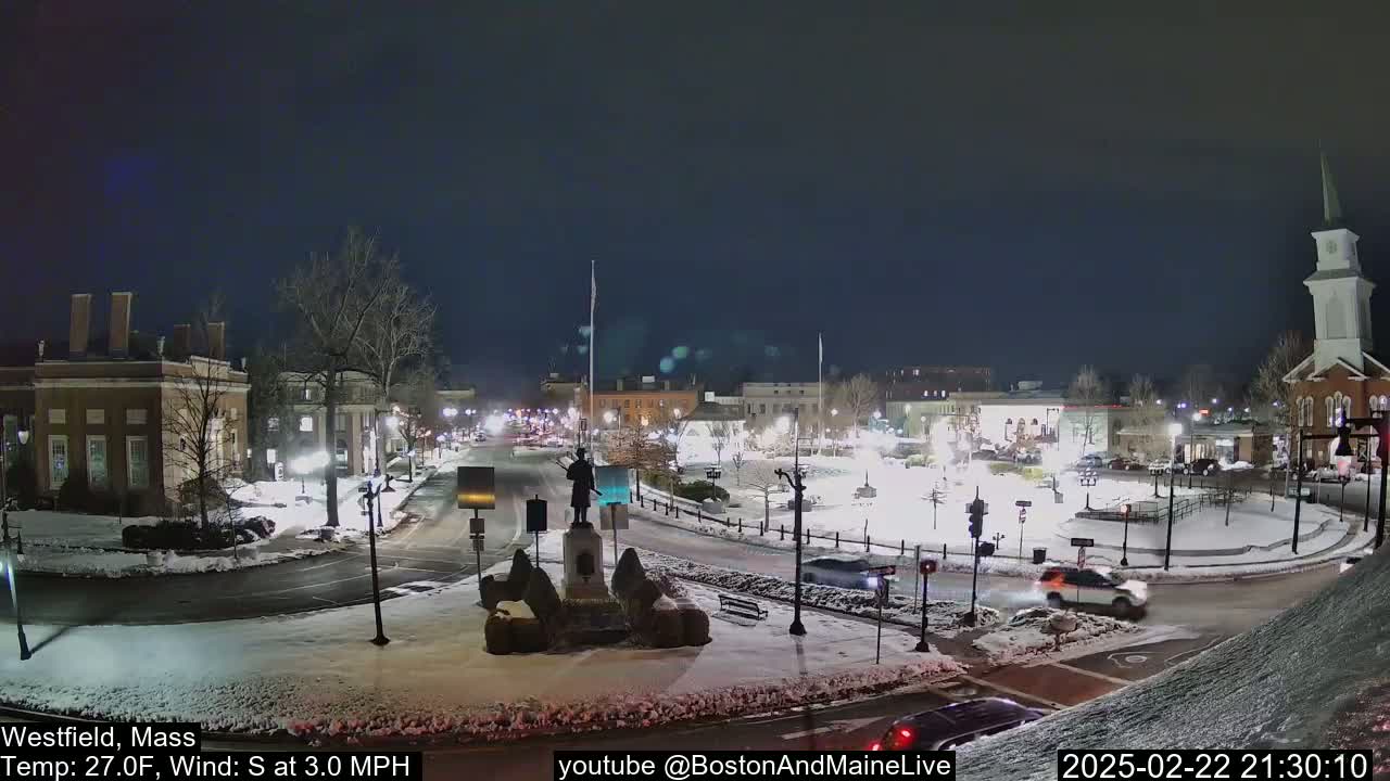A snow-covered town square at night, illuminated by streetlights, with a statue in the center and cars driving around it.