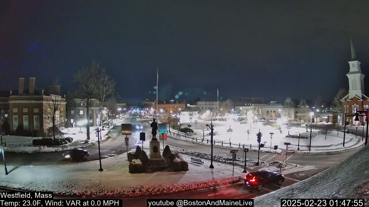 A snow-covered town square at night, illuminated by streetlights, features a statue in the center and several buildings surrounding it.
