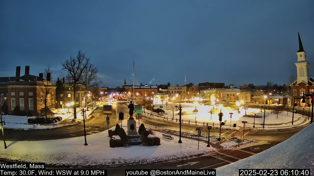 A snow-covered town square at night, illuminated by streetlights, features buildings, a statue, and a church steeple under a twilight sky.