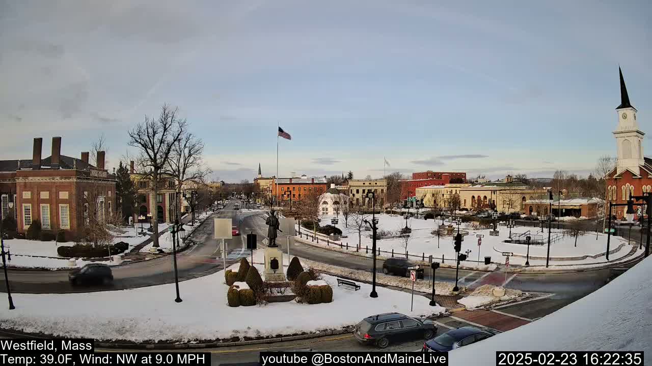 A snow-covered town square with brick buildings, a statue, and a church steeple under a partly cloudy sky.