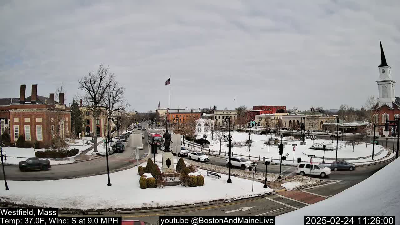 A snow-covered town square with several buildings, vehicles, and a monument, under a cloudy sky.