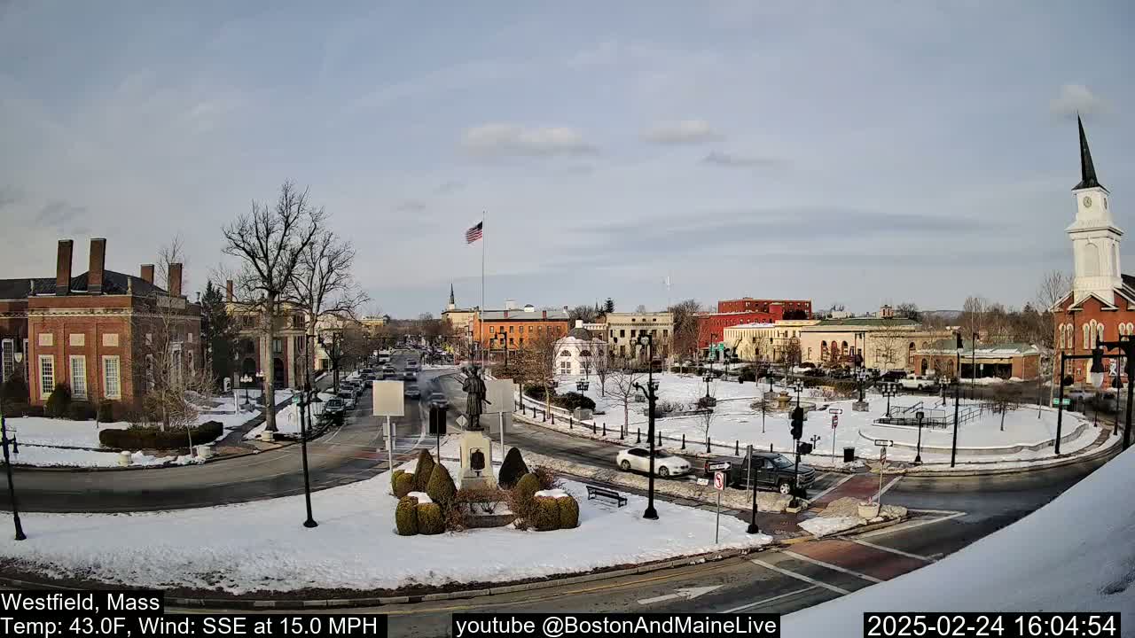 A snow-covered town square with brick buildings, a monument, and a church steeple under a partly cloudy sky.