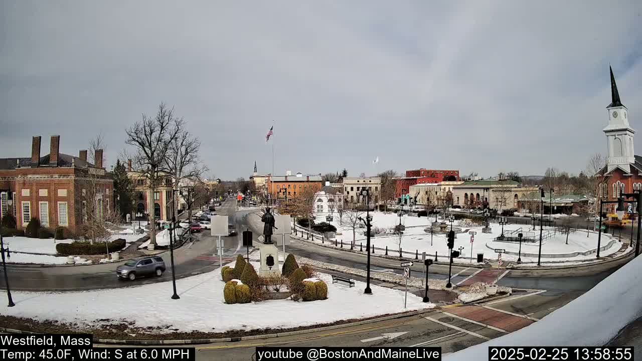 A snow-covered town square with brick buildings, a statue, and a church steeple under a cloudy sky.