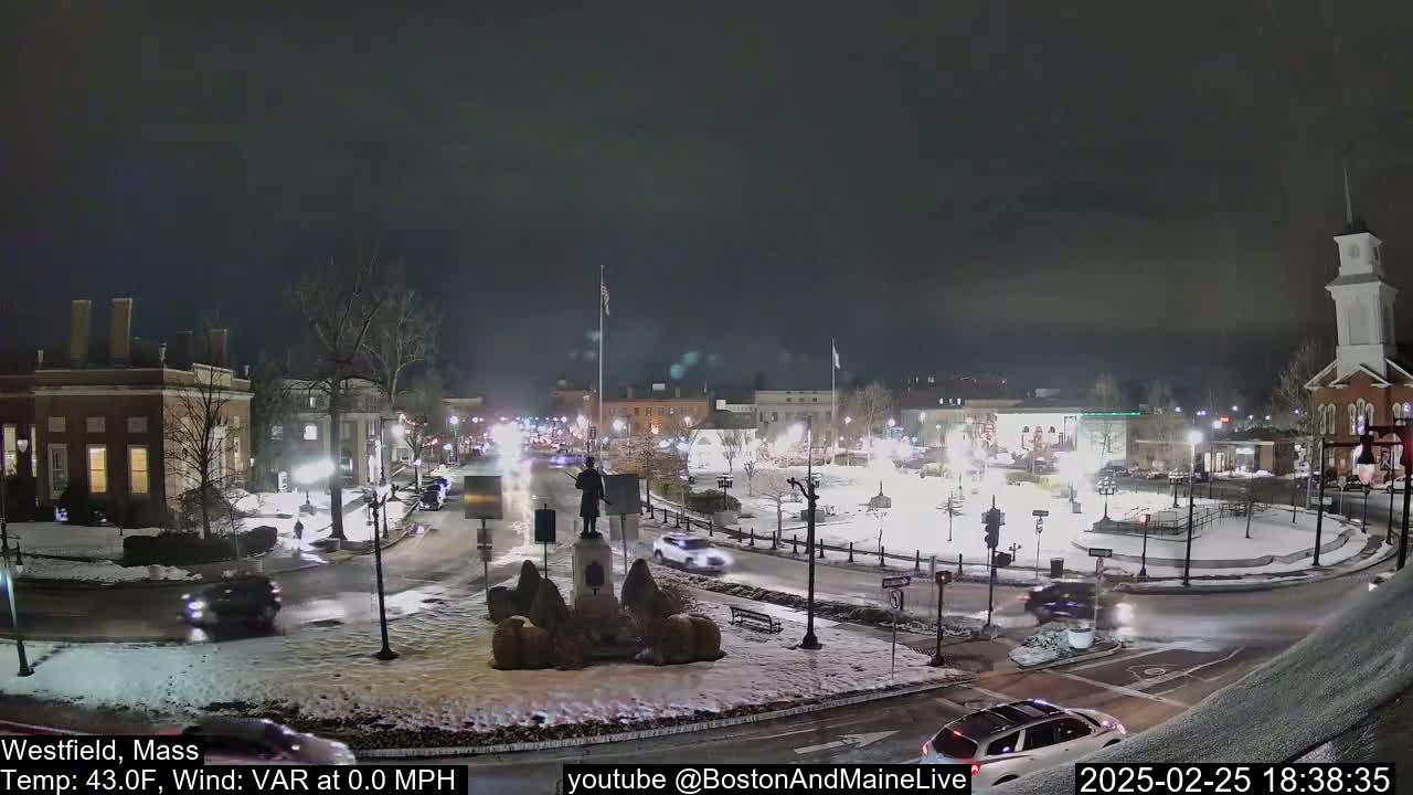 A snow-covered town square at night, illuminated by streetlights, with cars driving around a central monument and buildings surrounding the square.