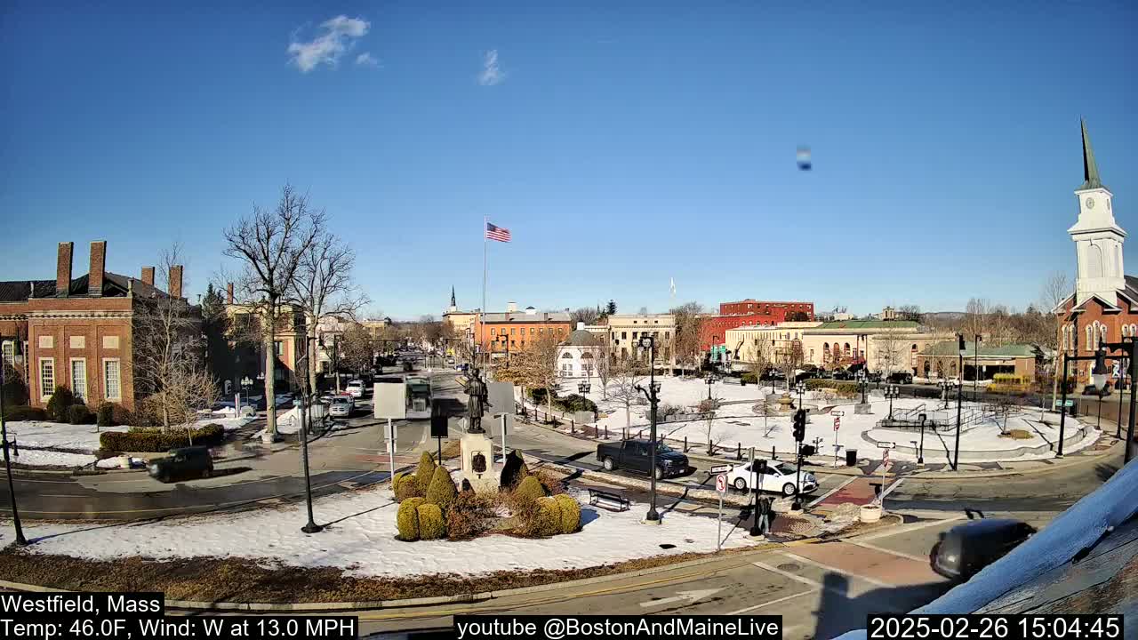 A town square with brick buildings, a few cars, and patchy snow on the ground under a clear blue sky.