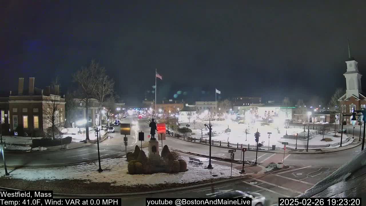 A snow-covered town square at night, illuminated by streetlights, features a statue in the center and several buildings surrounding it.