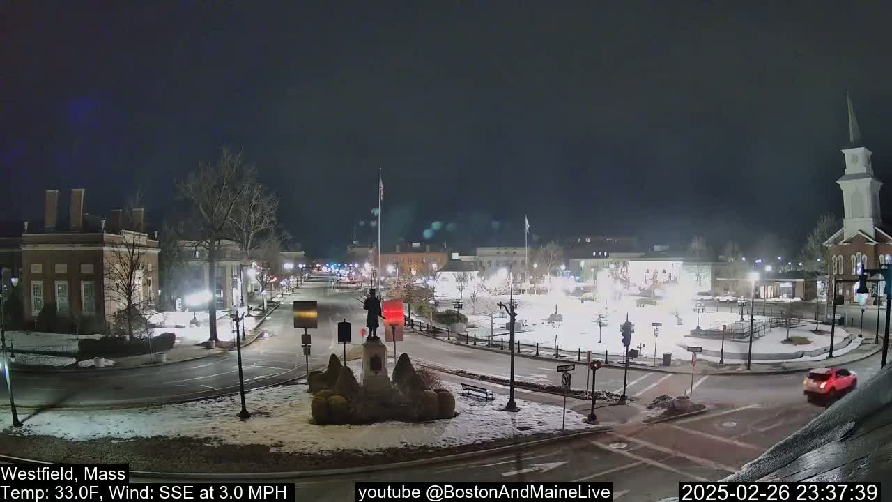 A snow-covered town square at night, illuminated by streetlights, features a statue in the center and several buildings surrounding it, with a calm wind blowing from the SSE at 3 mph and a temperature of 33°F.