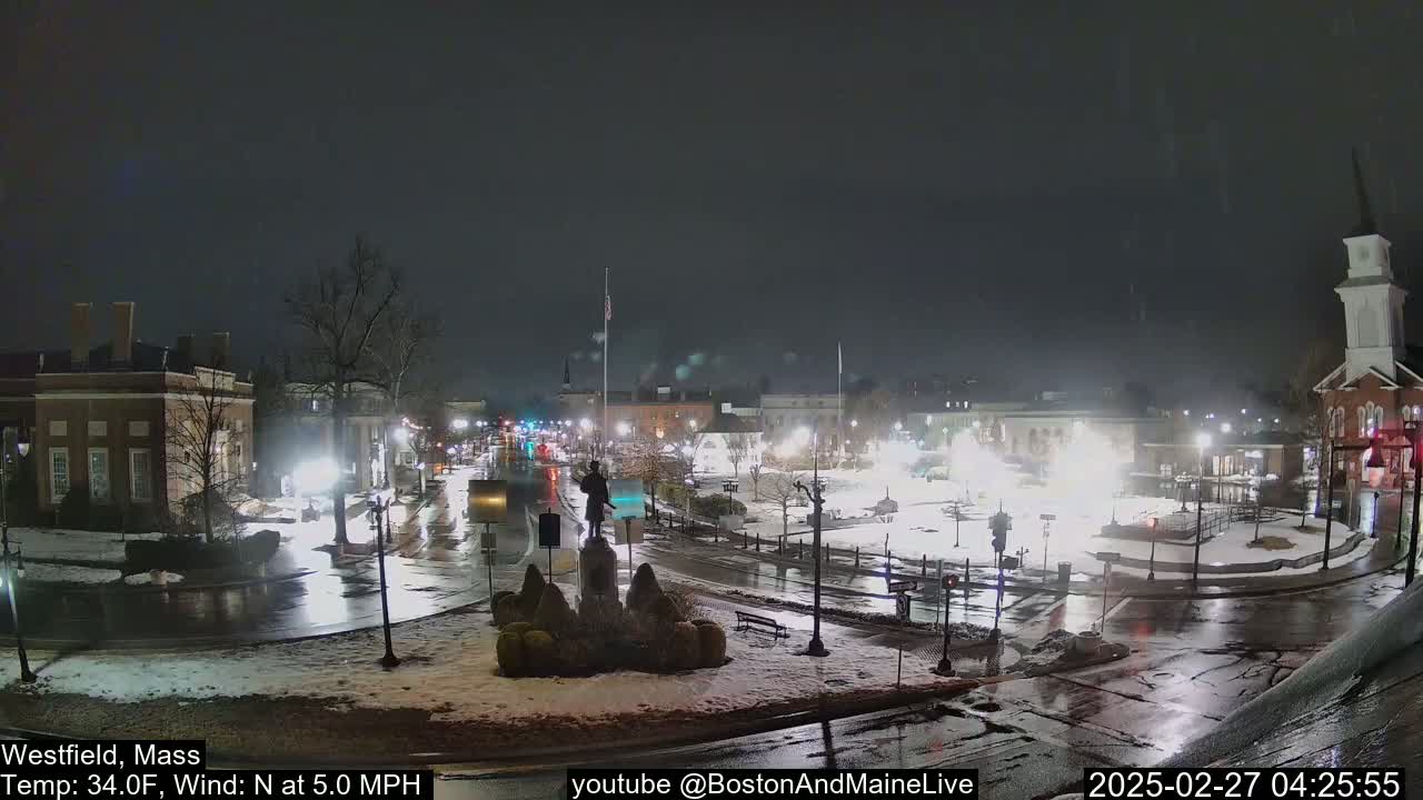 A nighttime view of a town square with wet roads and light snow cover, under a dark sky.