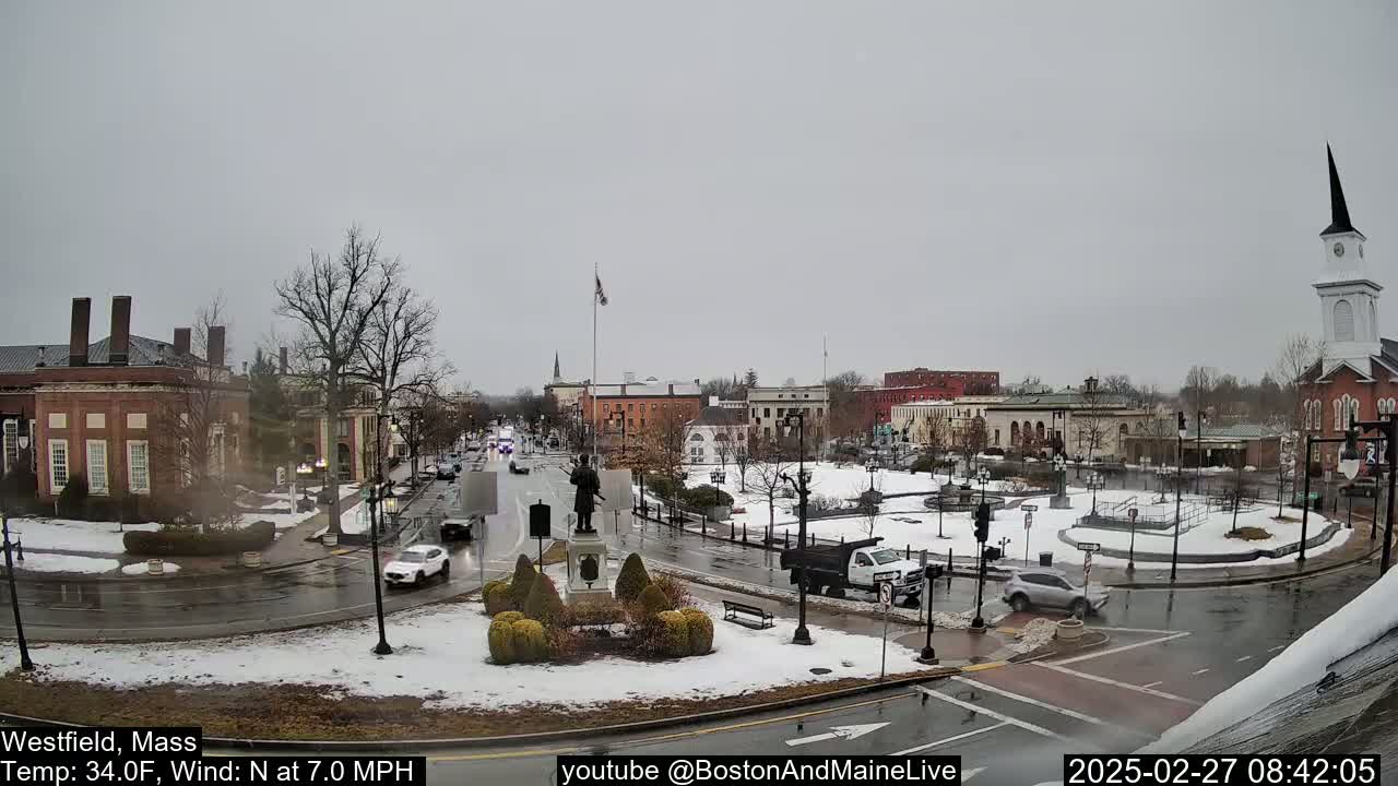 A partly snowy town square with several brick buildings, cars, and a statue in the center, under an overcast sky.