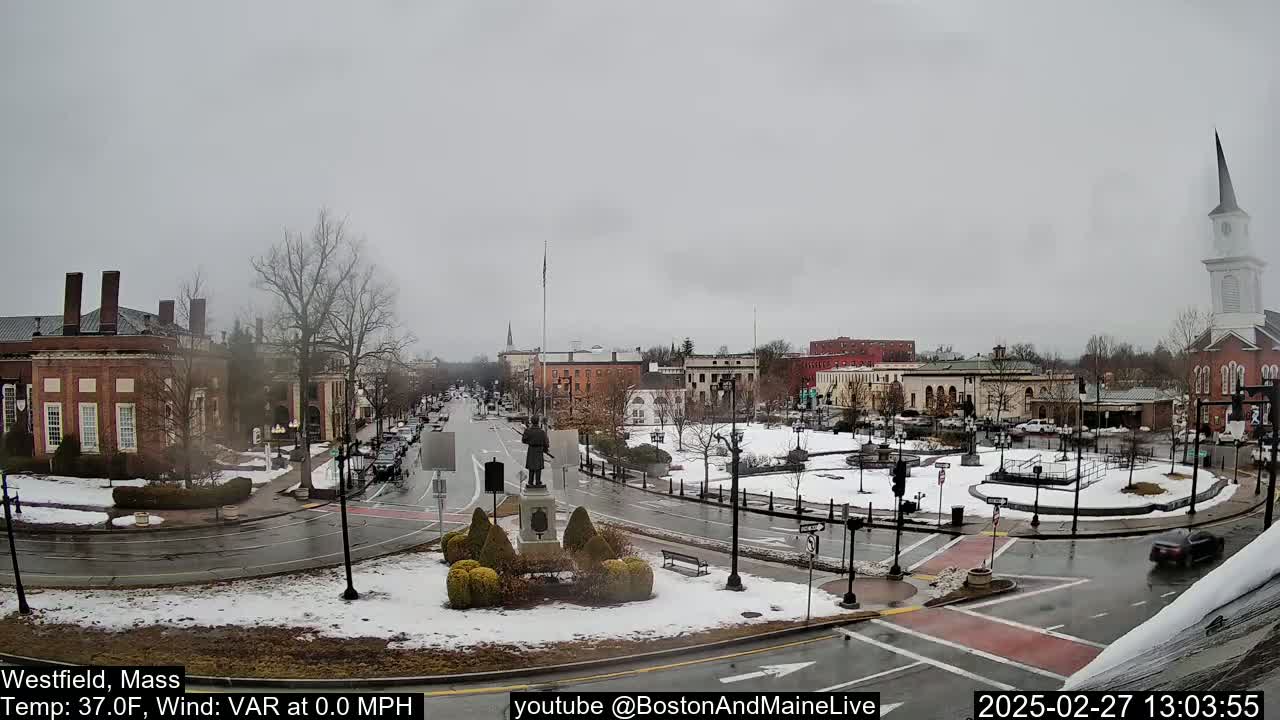 A snow-dusted town square, featuring brick buildings, a statue, and a church steeple, is visible under an overcast sky.