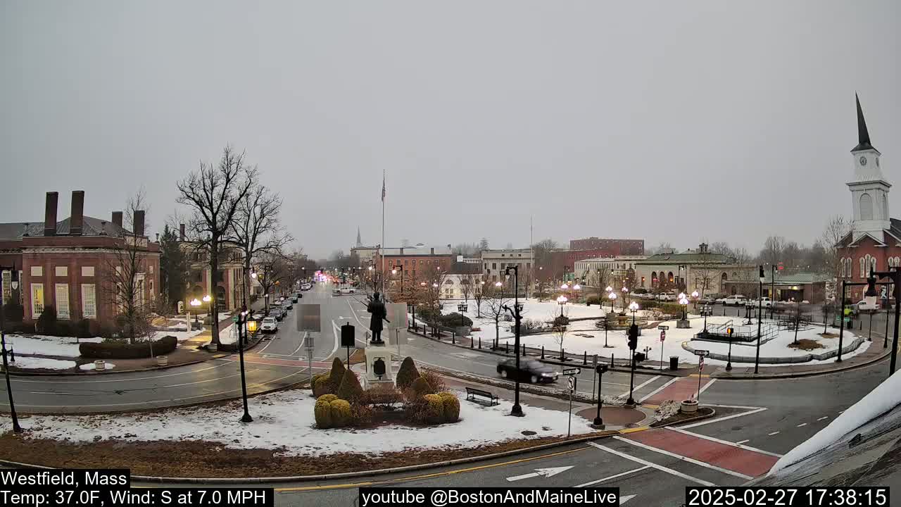 A town square with a statue and several brick buildings is partially covered in snow under an overcast sky.