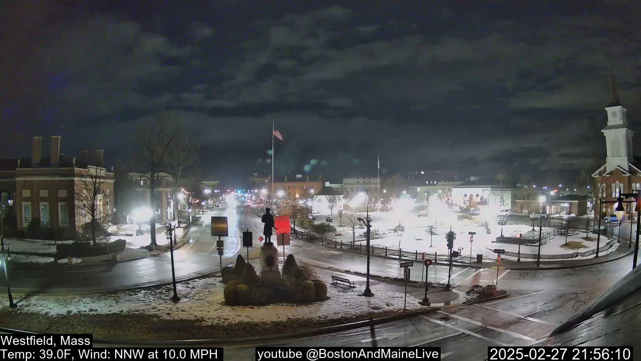 A nighttime view of a town square with a statue at its center, under a cloudy sky, with light snow on the ground and a temperature of 39 degrees Fahrenheit.
