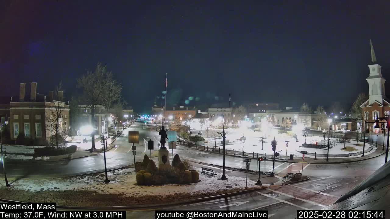 A nighttime view of a town square with snow-covered ground and buildings illuminated under a clear sky.