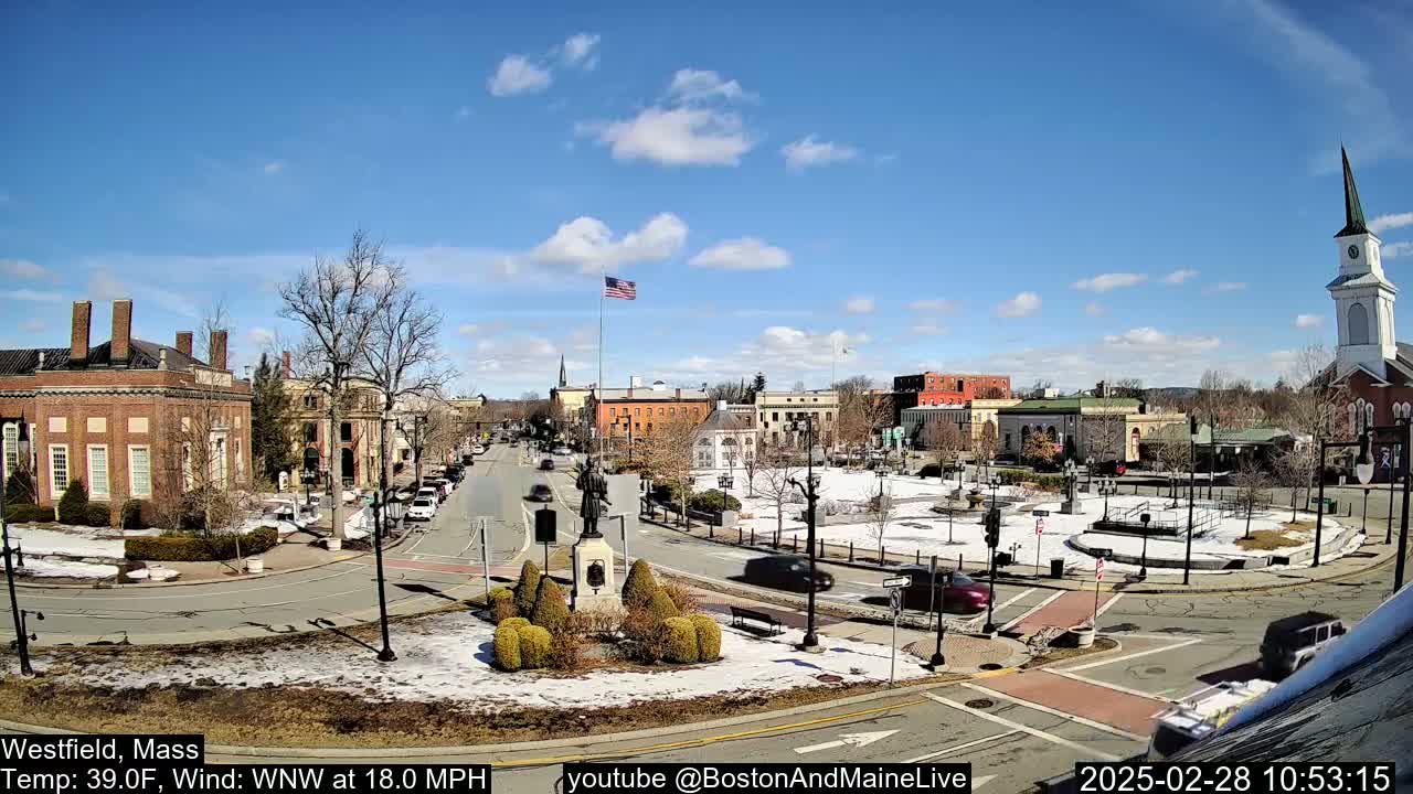 A town square with brick buildings and a church steeple under a mostly sunny sky with some clouds, and patches of snow on the ground.