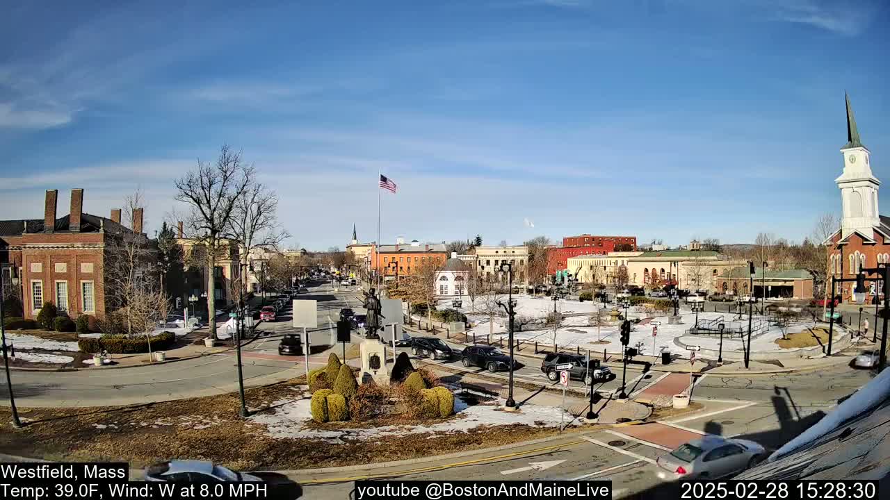 A town square with brick buildings, a statue, and sparse traffic under a clear, sunny sky.