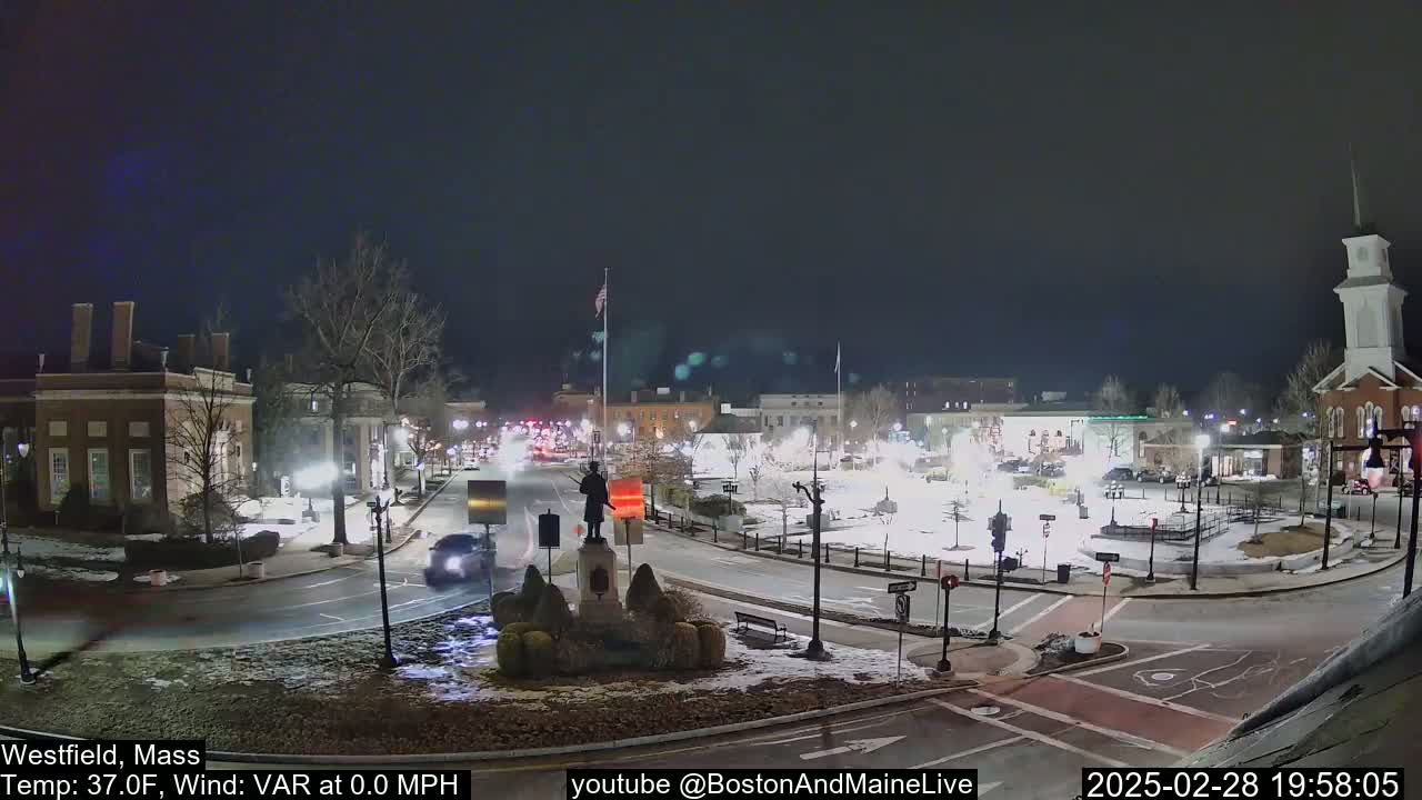 A nighttime view of a town square with snow on the ground, featuring a statue in the center and several buildings surrounding it under a dark sky.