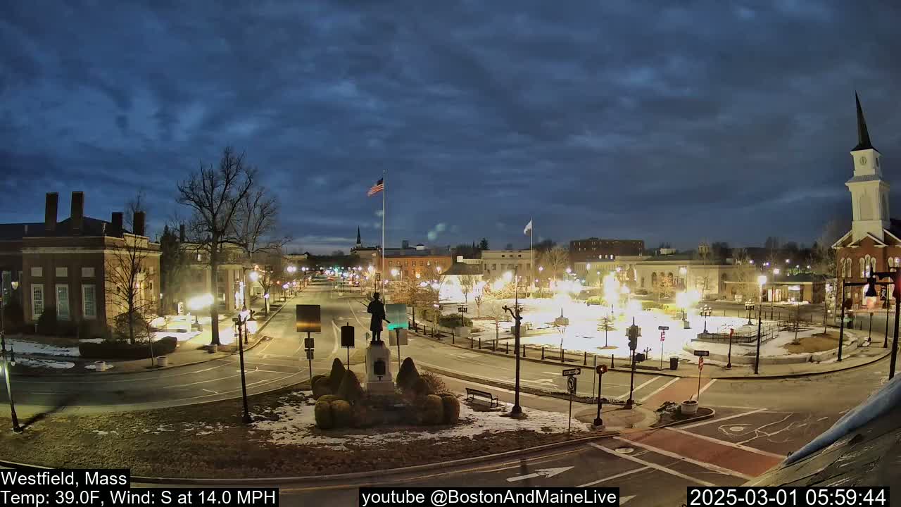 A mostly clear, twilight sky over a town square at night, with snow on the ground and streetlights illuminating the area, including a statue in the center.