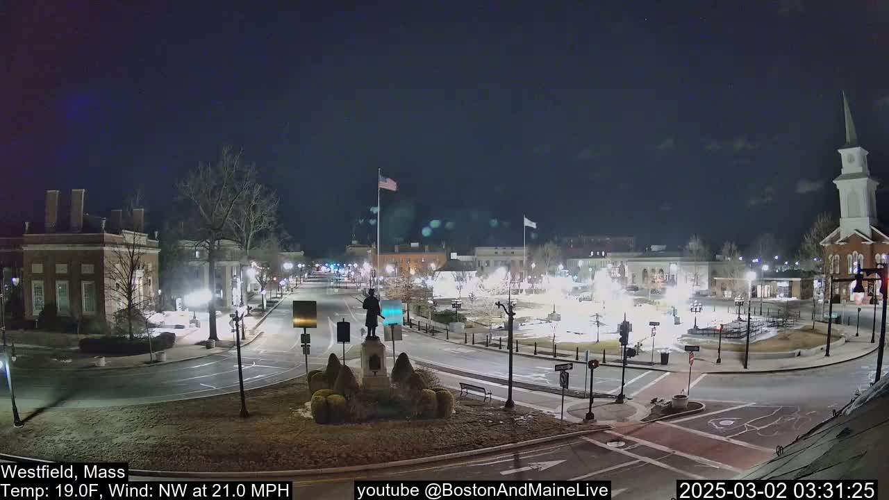 A nighttime view of a town square with a statue, buildings, and roads, under a clear, dark sky with a northwest wind.