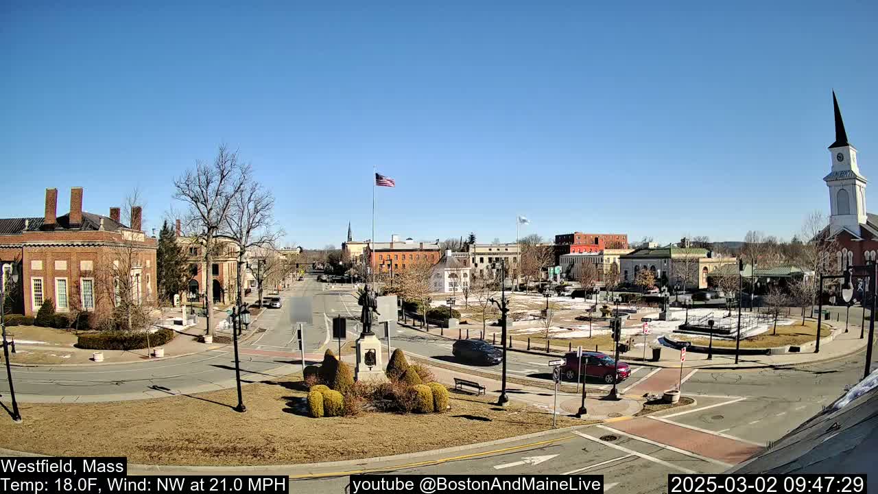 A town square with brick buildings, a statue, and sparse traffic under a clear blue sky.