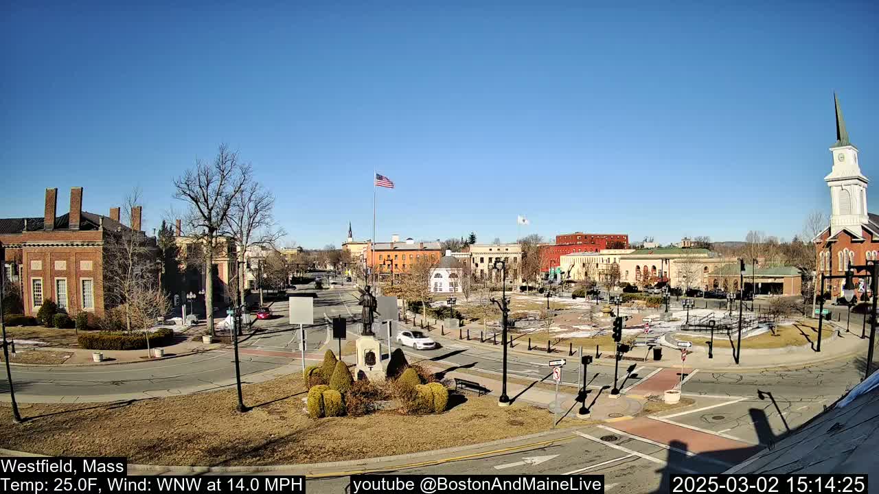 A town square with brick buildings, a monument, and a church steeple under a clear blue sky on a sunny, windy day.