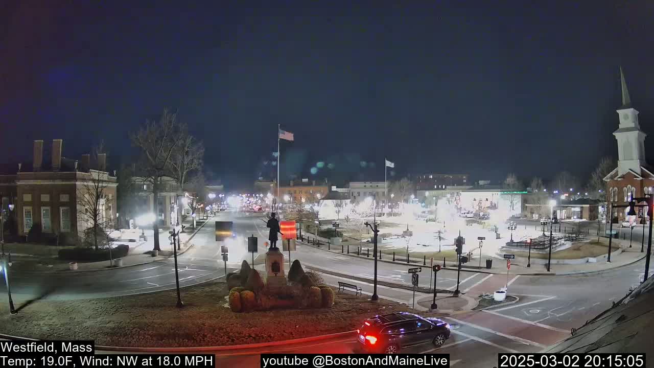 A nighttime, low-temperature view of a town square with a statue at its center, surrounded by buildings and cars under a mostly clear sky, with a northwest wind at 18 mph.