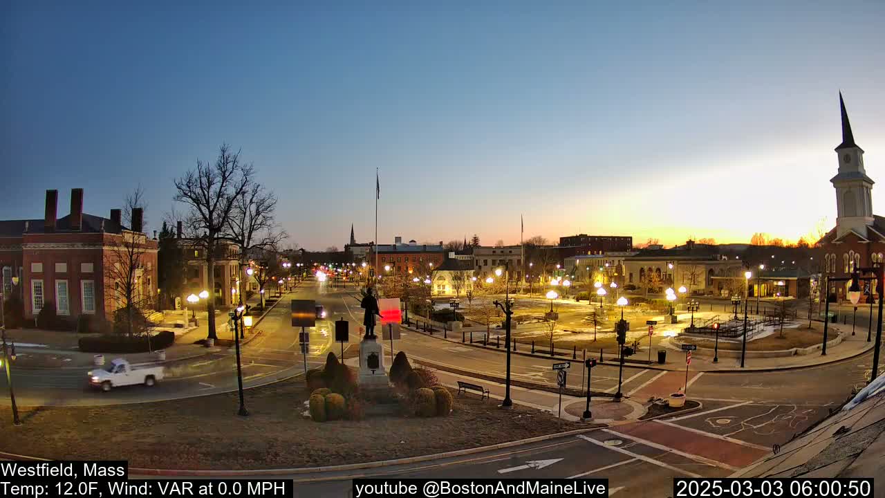 A town square at twilight, under a clear sky, is illuminated by streetlights, with a statue at its center and a church steeple in the background.
