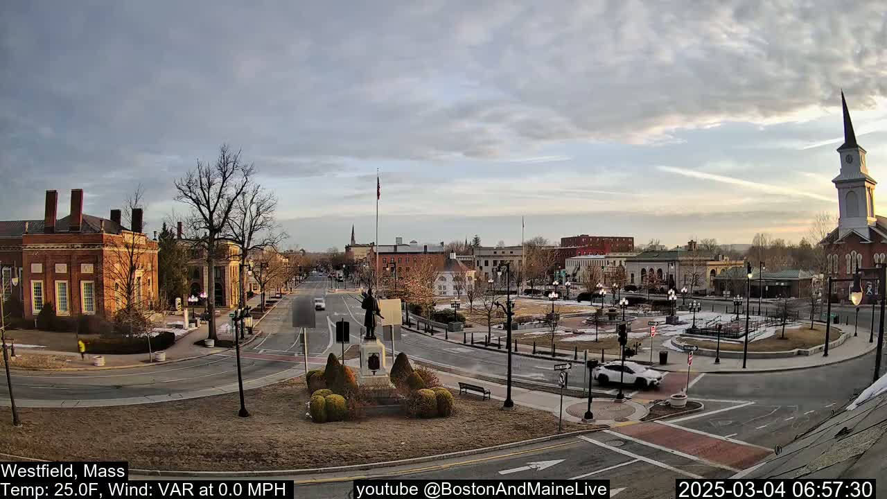 A mostly cloudy, cool morning reveals a town square with brick buildings, a statue, and a church steeple, with a few cars on the roads.
