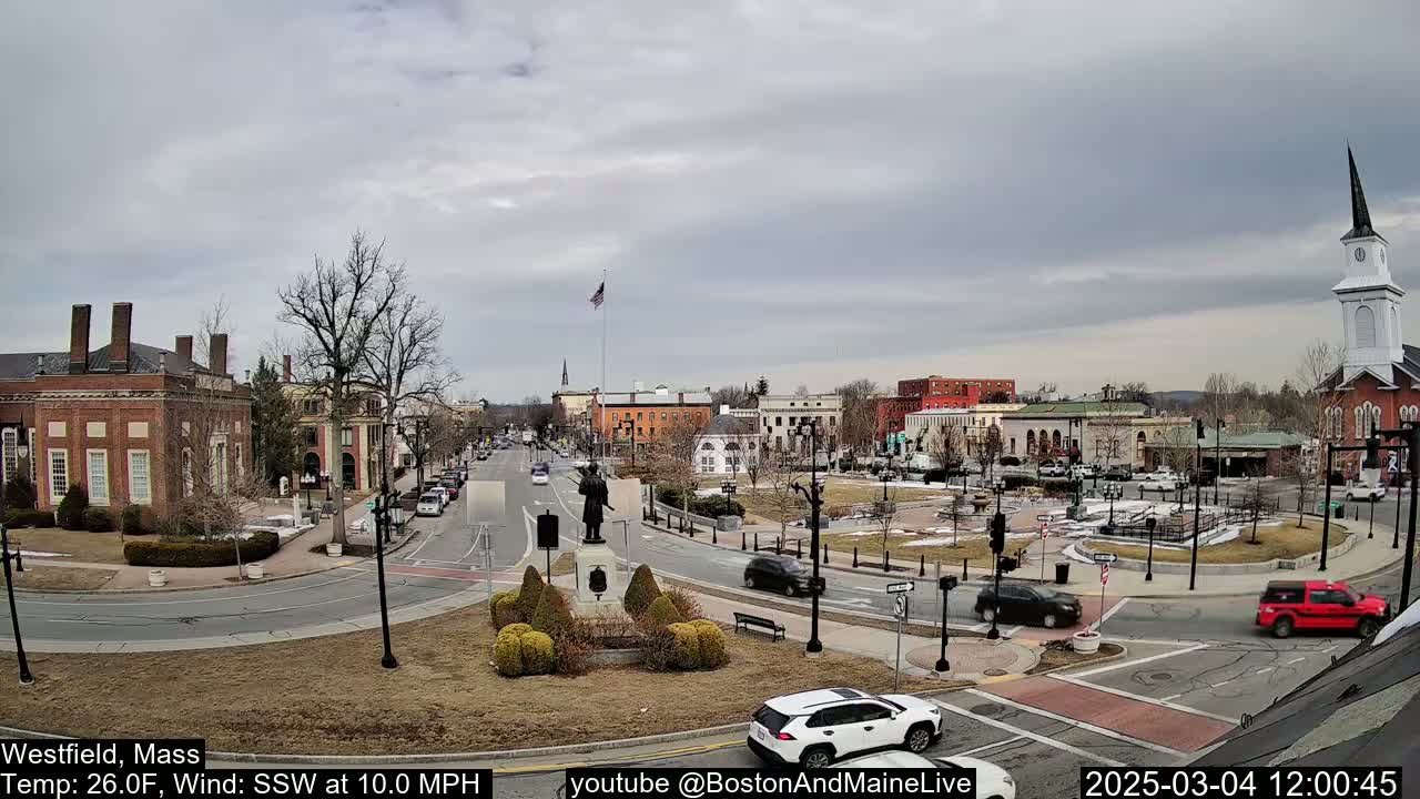 A town square with brick buildings, a statue, and sparse traffic under an overcast sky.