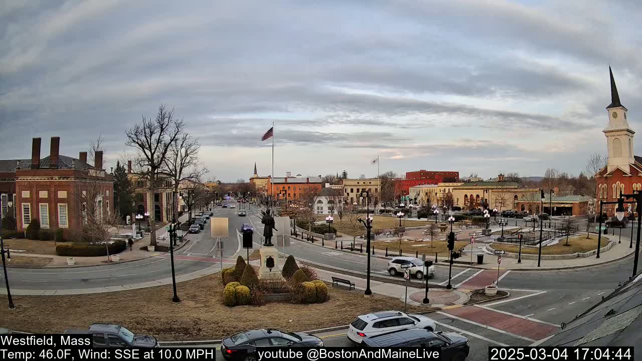 A town square with brick buildings, a statue, and sparse traffic under a cloudy sky.