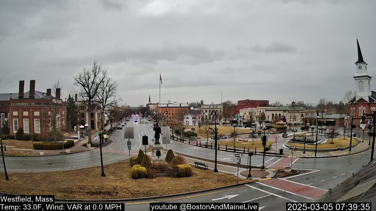 A town square with a statue in the center, surrounded by brick buildings and a church steeple, under an overcast sky.