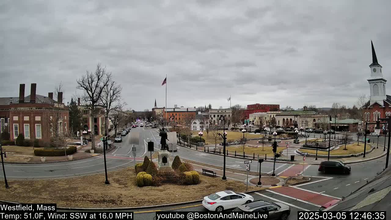 A town square with brick buildings, a monument, and sparse traffic under an overcast sky.