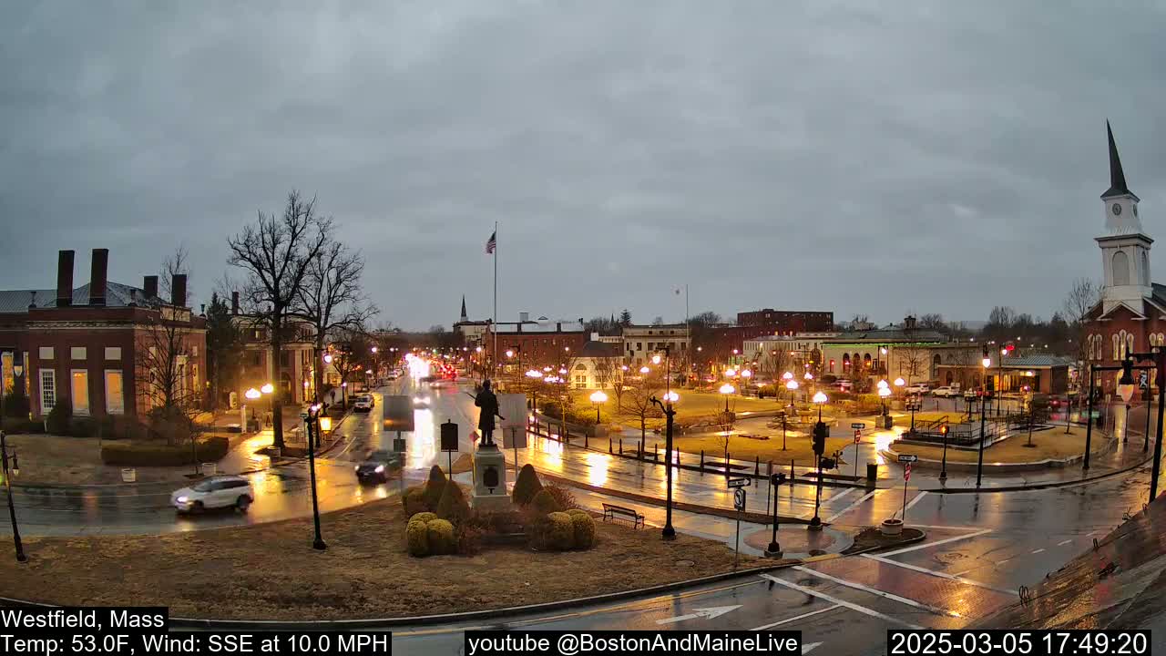 A rain-slicked town square at dusk, featuring brick buildings, streetlights, and a statue in a small park, with a church steeple in the background.
