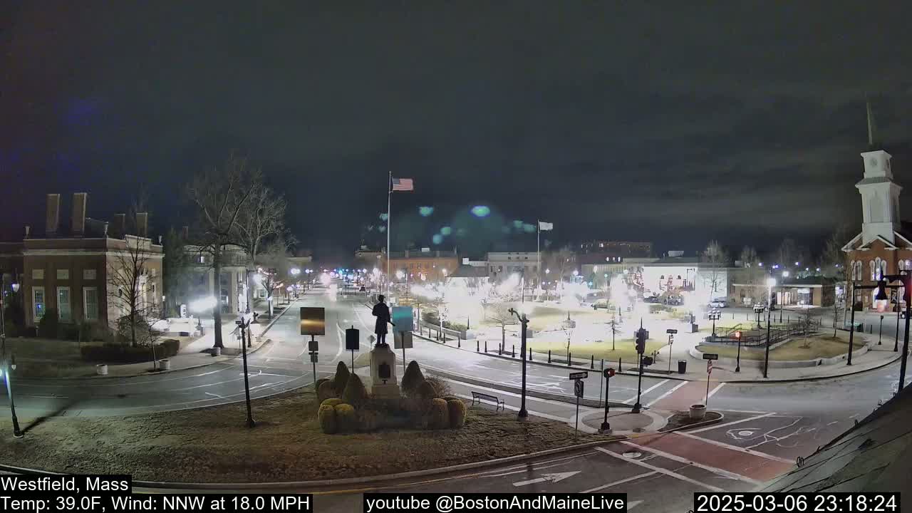 A nighttime, low-wind view of a town square with a statue at its center, illuminated buildings surrounding it, and a few cars on the roads.