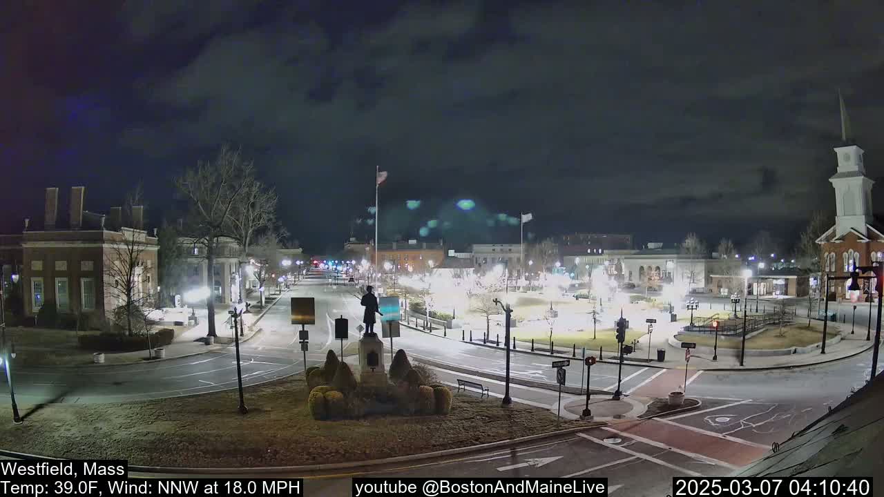 A nighttime view of a town square with a statue in the center, under a mostly cloudy sky.