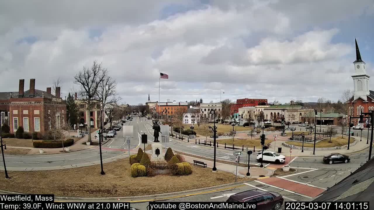 A town square with brick buildings, a monument, and sparse landscaping under a cloudy sky.