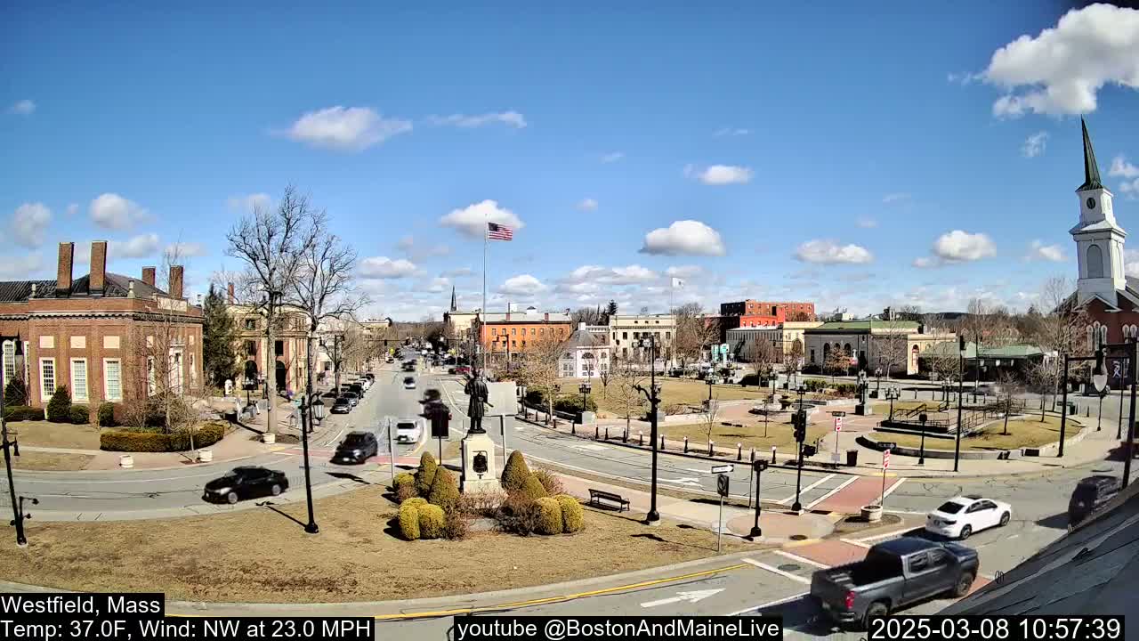 A sunny town square with brick buildings, a statue, and sparse traffic under a partly cloudy sky.