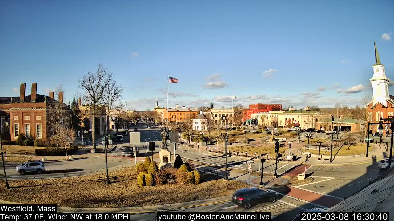A sunny town square with brick buildings, a church steeple, and sparse traffic under a mostly clear blue sky.