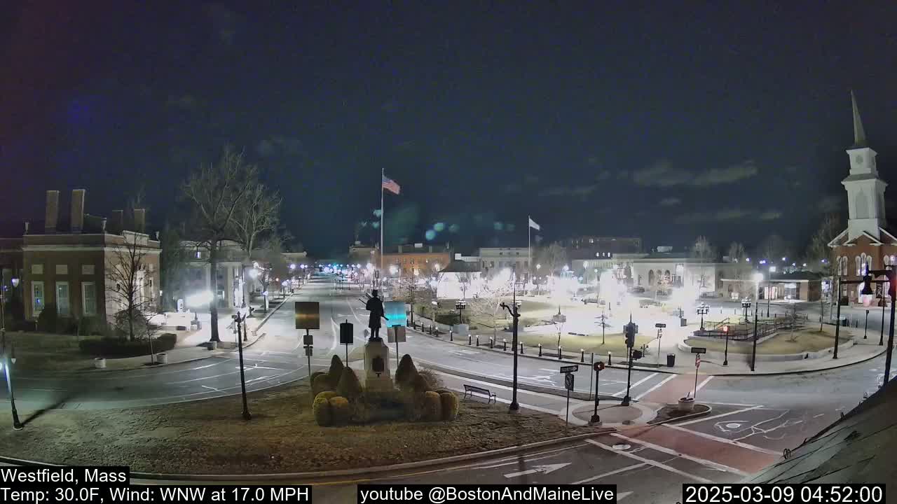 A nighttime view of a town square with brick buildings, a statue, and a church steeple, under a clear, dark sky with a 30°F temperature and a 17 mph wind from the west-northwest.