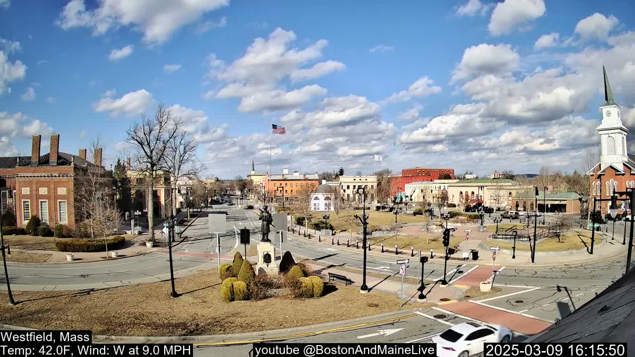 A partly cloudy day reveals a town square with brick buildings, a monument, and a church steeple, surrounded by roads and sparse trees.