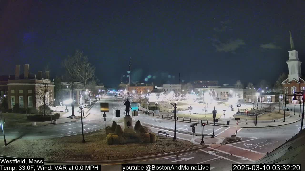 A nighttime view of a town square with a statue at its center, surrounded by buildings and relatively empty streets under a mostly clear sky.