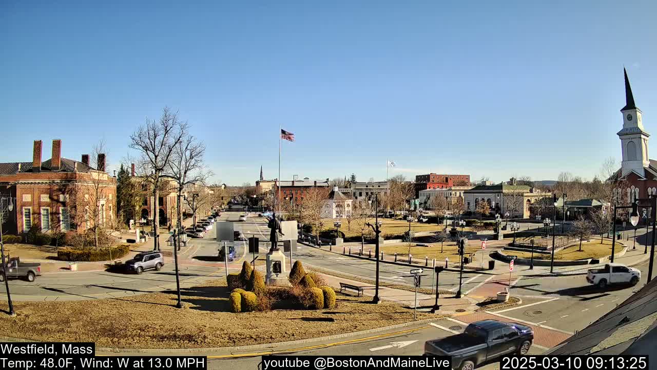 A sunny town square with brick buildings, a statue, and sparse, leafless trees, under a clear blue sky.