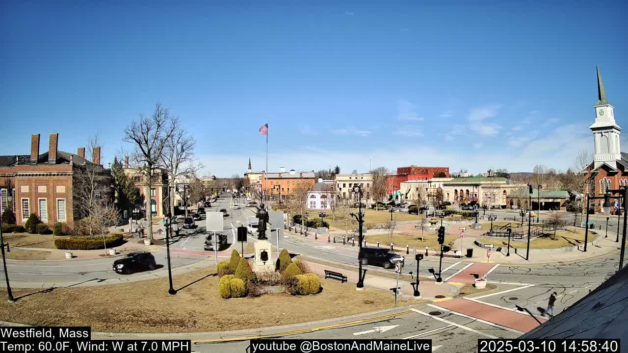 A town square with brick buildings, a monument, and sparse traffic under a clear blue sky.