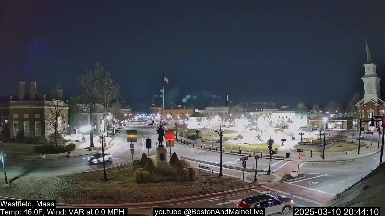 A nighttime view of a town square with a statue at its center, surrounded by buildings and sparse traffic under a clear, dark sky.