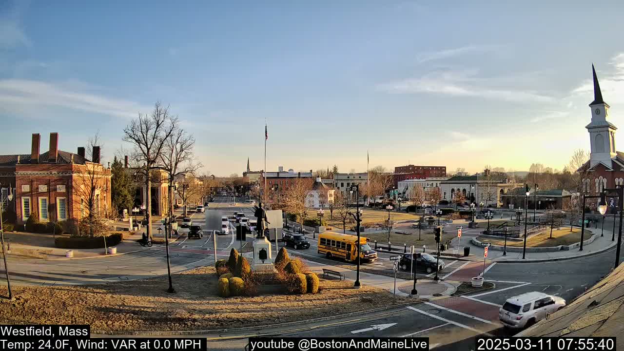 A town square with brick buildings, a statue, a school bus, and several cars is visible under a clear, sunny sky.