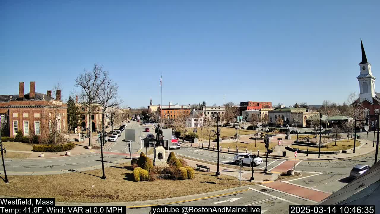 A town square with brick buildings, a statue, and sparse traffic under a clear blue sky.