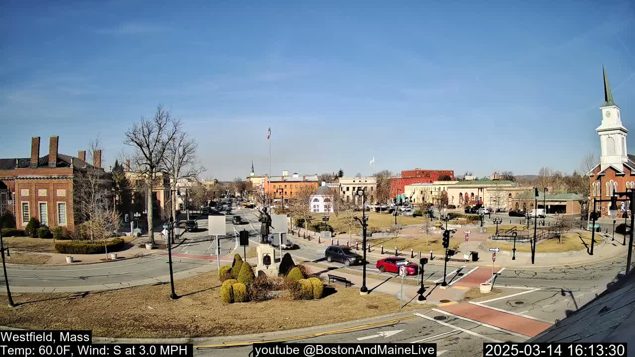 A sunny town square with brick buildings, a monument, and sparse traffic under a clear blue sky.