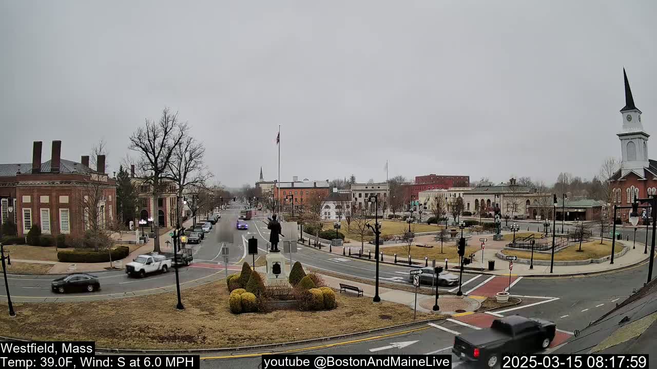 A town square with brick buildings, a statue, and sparse traffic under an overcast sky.
