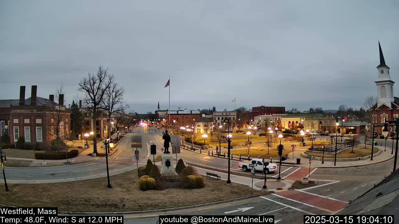 A town square at dusk under a cloudy sky, featuring a statue in the center, surrounded by buildings and streetlights.