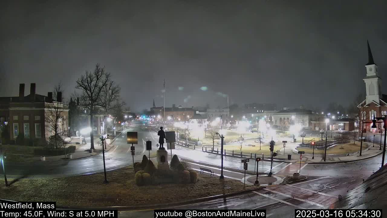A nighttime view of a town square with a statue at its center, under a dark, misty sky with a slight south wind.