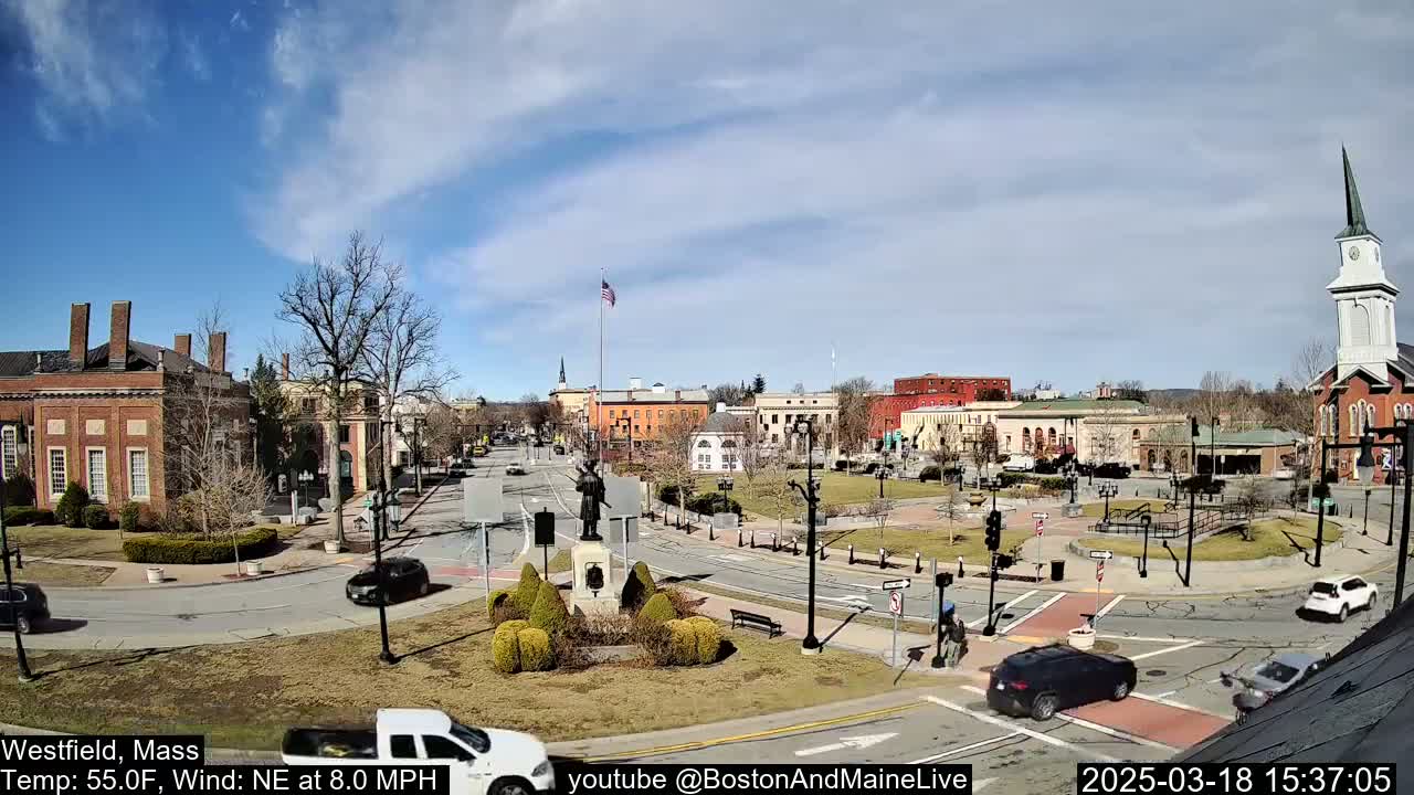 A town square with brick buildings, a monument, and sparse traffic under a partly sunny sky.